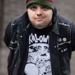 A photo of Brian Transplant wearing a black denimn vest and a green-and-red striped hat.